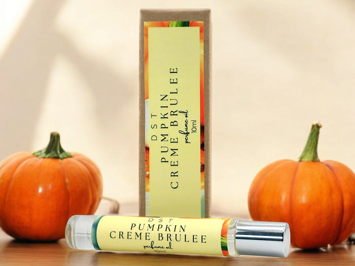 10ml bottle of Pumpkin Creme Brulee perfume oil and the box it is packaged in sitting on wood countertop with 2 pumpkins and cream-colored background.