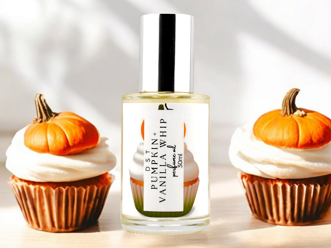 30 milliliter bottle of pumpkin and vanilla whip perfume oil sitting on a light wood countertop with 2 vanilla cupcakes with orange frosting and orange fondant pumpkins on top against a shadowy white background.