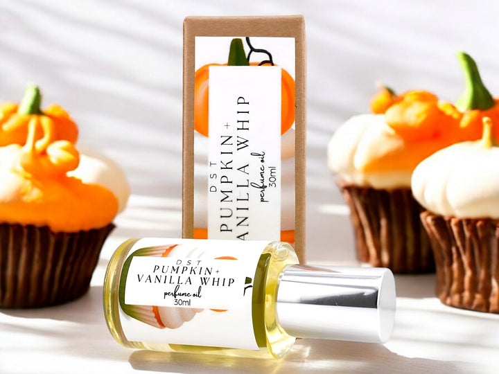 30 milliliter bottle of pumpkin and vanilla whip perfume oil and the box that it is packaged in sitting on a light wood countertop with three vanilla cupcakes with orange frosting and orange fondant pumpkins on top against a shadowy white background.