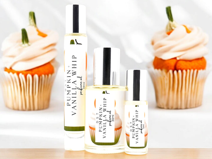 5, 10, and 30 milliliter bottles of pumpkin and vanilla whip perfume oil and the box that it is packaged in sitting on a light wood countertop with 2 vanilla cupcakes with orange frosting and orange fondant pumpkins on top against a shadowy white background.