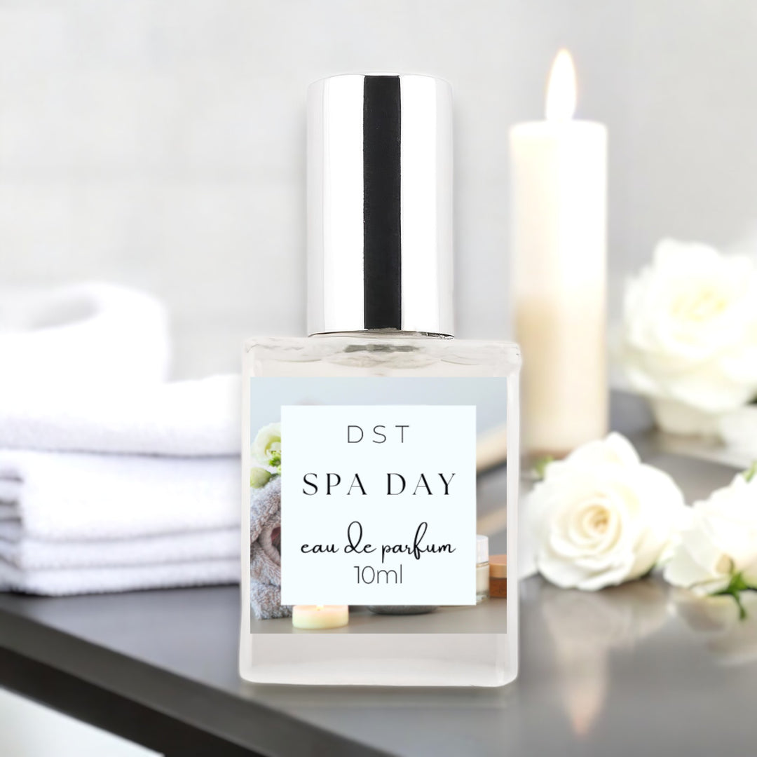 10 milliliter bottle of spa day perfume sitting on a gray countertop with white towels, a white lip candle, and a bouquet of white roses.