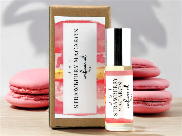 5ml bottle of Strawberry macaron perfume oil and the box it is packaged in sitting on wood countertop with pink macarons stacked against a white shadowy background.