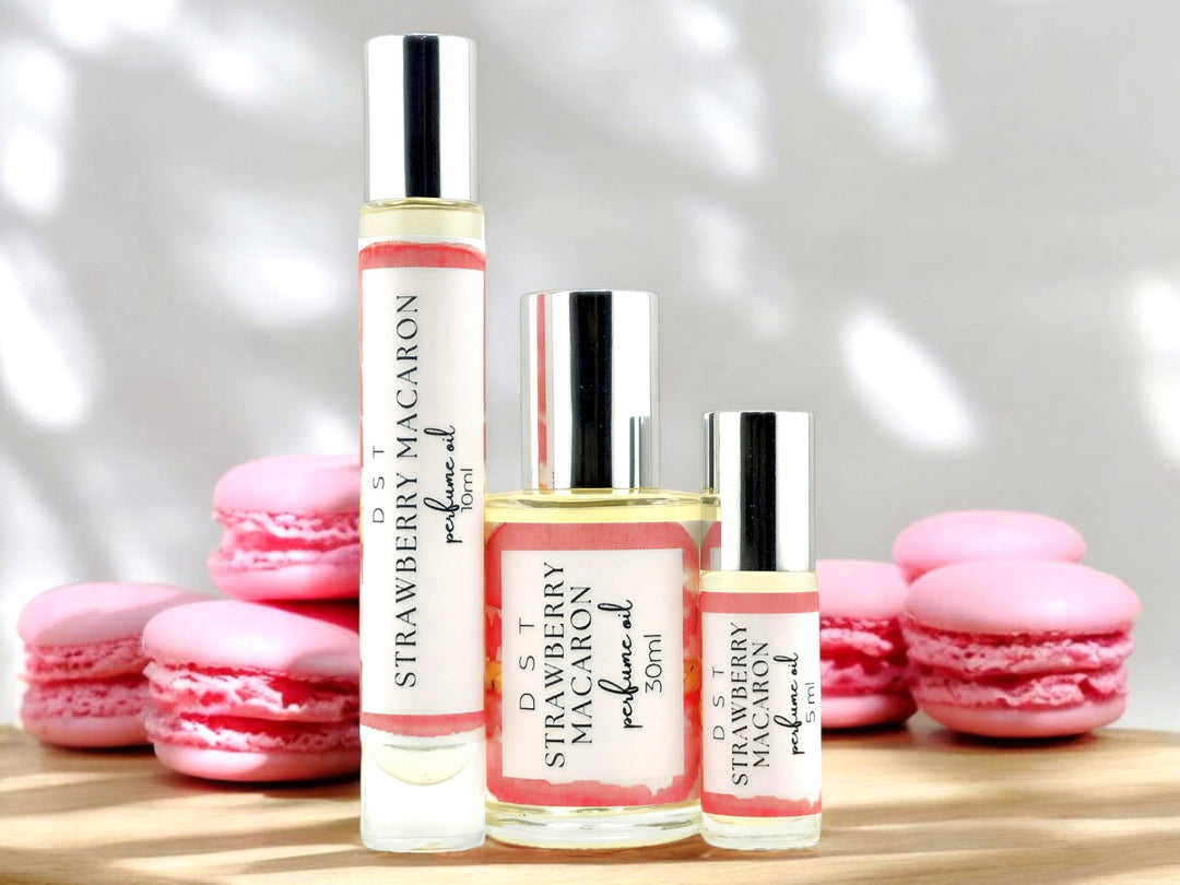 5, 10 and 30ml bottles of Strawberry macaron perfume oil sitting on wood countertop with pink macarons stacked against a white shadowy background.