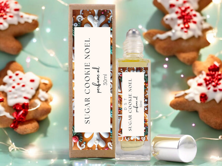 30ml bottle of Sugar Cookie Noel Perfume Oil and the box it is packaged in sitting on a glass surface with decorated gingerbread cookies and twinkling fairy lights against pastel green background.