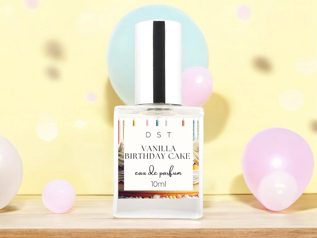 10ml bottle of Vanilla birthday Cake Eau de Parfum spray sitting on a wood countertop with pastel pink and blue balloons against a pastel yellow background.