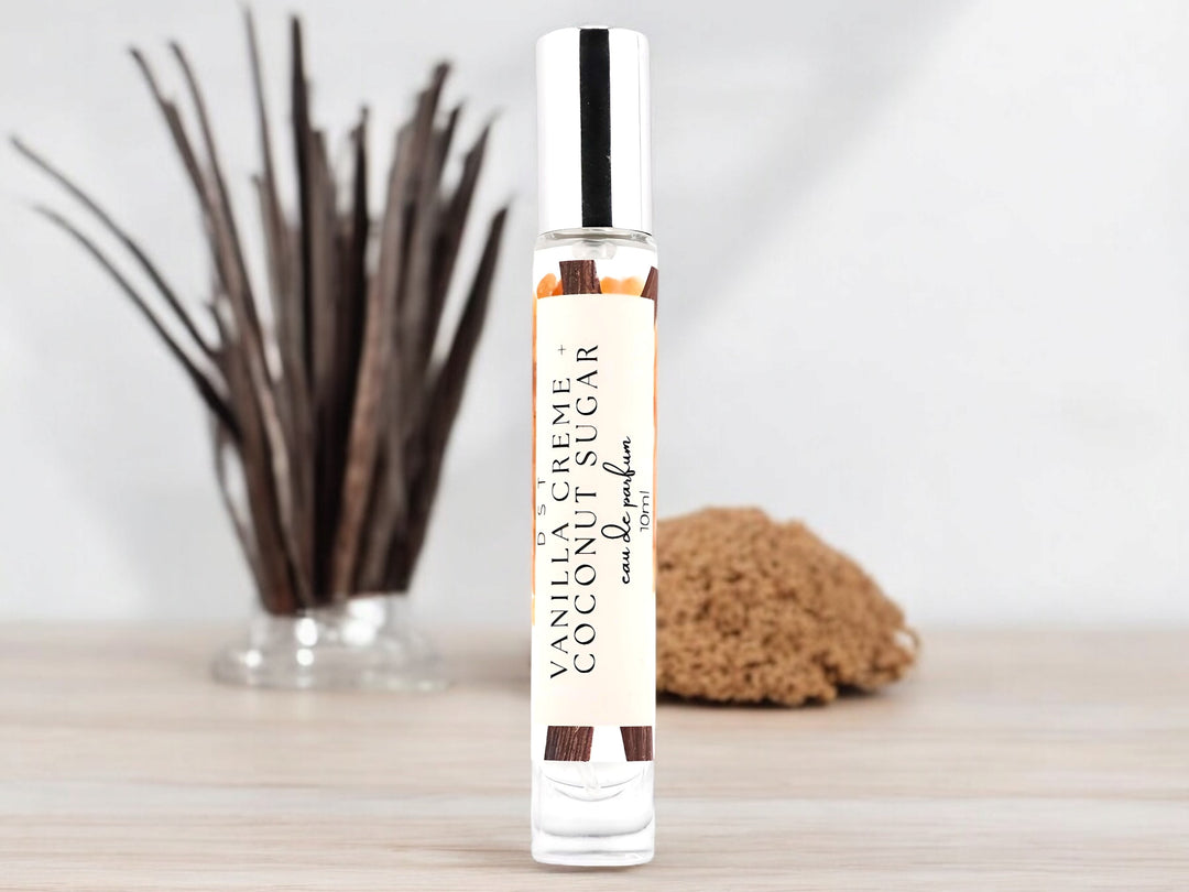 10ml bottle of Vanilla Creme and Coconut Sugar Eau de Parfum sitting on a wood countertop with a chunk of brown sugar and long vanilla beans sticking out of a glass vase against a white background.