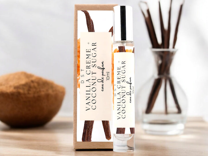 10ml bottle of Vanilla Creme and Coconut Sugar Eau de Parfum and the box it is packaged in sitting on a wood countertop with a chunk of brown sugar and long vanilla beans sticking out of a glass vase against a white background.