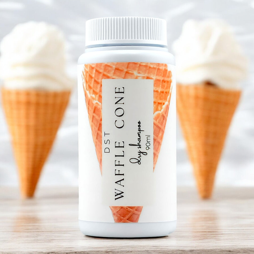 90ml bottle of Waffle Cone scented Dry Shampoo powder sitting on a wood countertop with 2 vanilla Waffle Cones in the background.