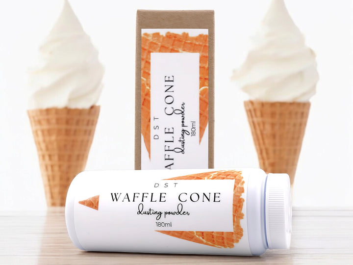 A 180ml bottle of waffle cone scented body powder and the box it is packaged in sitting on a wood countertop with 2 vanilla ice cream cones against a white background.