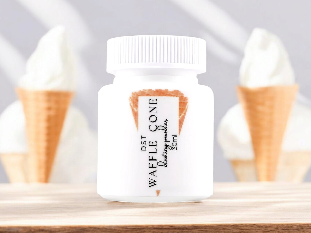 A 30ml bottle of waffle cone scented body powder sitting on a wood countertop with 2 vanilla ice cream cones against a white background.