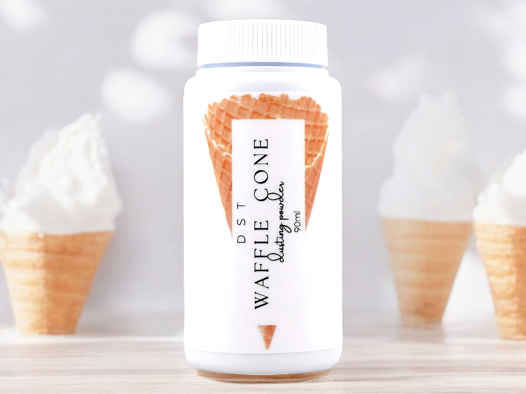 A 90ml bottle of waffle cone scented body powder sitting on a wood countertop with 3 vanilla ice cream cones against a white background.