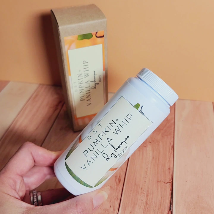 180ml bottle of Pumpkin and Vanilla Whip scented Dry Shampoo powder and the box it is packaged in sitting on wood counter top with light orange backdrop. 