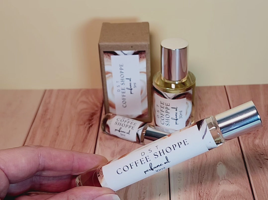 5, 10, and 30ml bottles of Coffee Shoppe Perfume oil and the box it is packaged in sitting on wood countertop with ivory background.