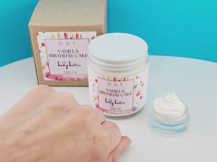 120ml jar of Vanilla Birthday Cake scented Body Butter and the box it is packaged in. Model shown rubbing a small amount of body butter on her hand to show consistency of product.