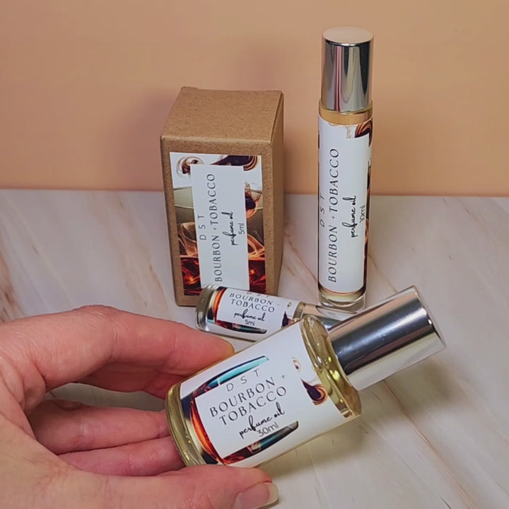 5, 10, thirty milliliter bottles of bourbon and tobacco perfume oil and the box it is packaged in sitting on a cream marble countertop with peach colored background.