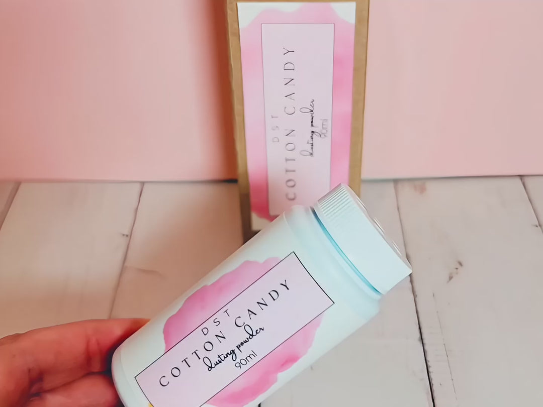 A 90ml bottle of Cotton Candy scented body powder and the box it is packaged in sitting on wood countertop with pale pink background.