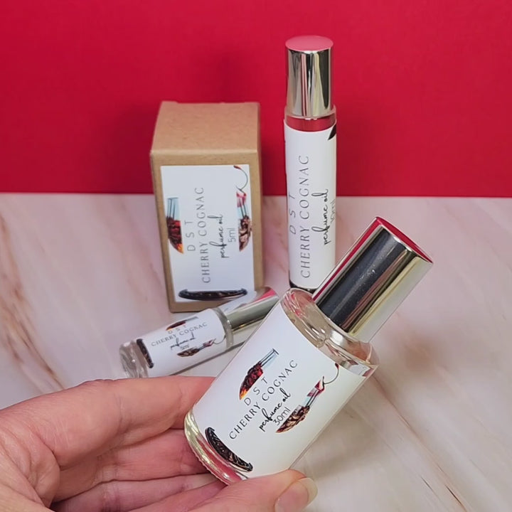 Five, 10, 30 milliliter bottles of Cherry Cognac perfume oil sitting on a cream marble countertop with bright red background.