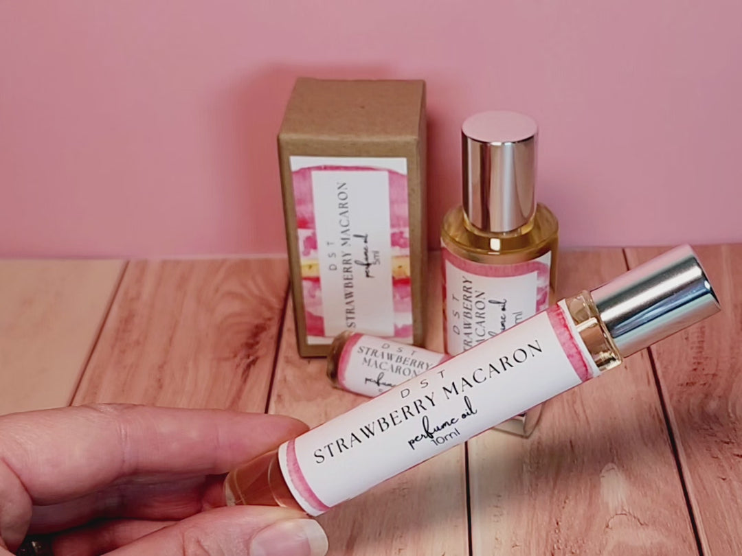 5, 10, and 30ml bottles of Strawberry Macaron perfume oil and the box it is packaged in sitting on wood countertop with pastel pink background.