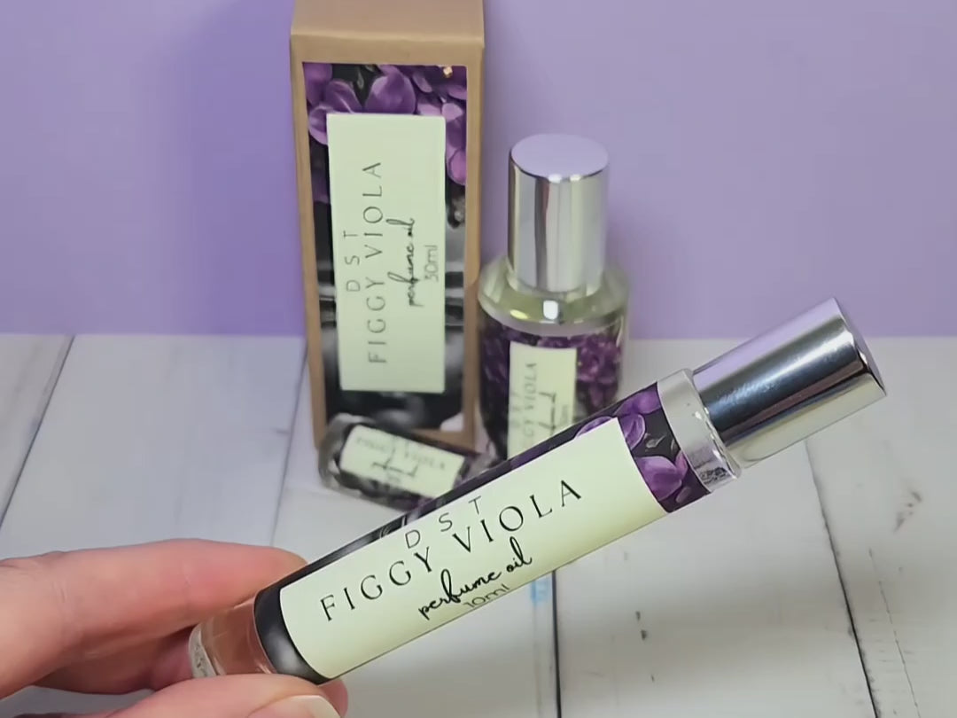 5, 10, and 30ml bottles of Figgy Viola Perfume Oil and the box that it is packaged in sitting on white wood countertop and lilac colored background.