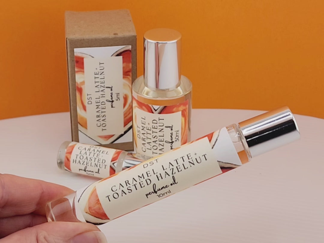 5, 10, and 30ml bottle of Caramel Latte and Toasted Hazelnut Perfume Oil sitting on white countertop with orange background. 