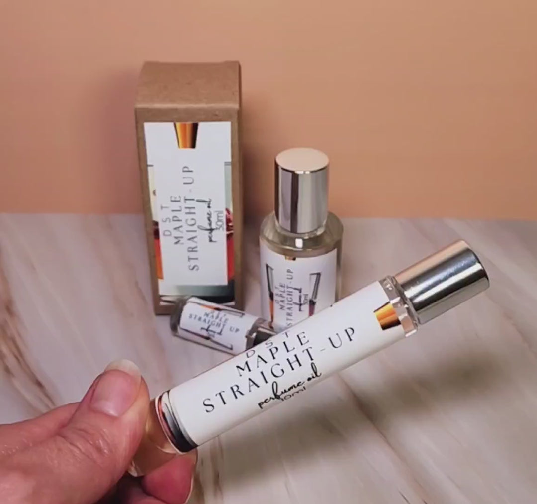 Five 10 and thirty milliliter bottles of Maple straight up perfume oil and the box that it is packaged in sitting on a tan marble countertop with peach background.