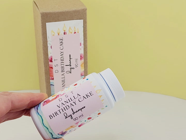 90ml bottle of Vanilla Birthday Cake scented Dry Shampoo powder and the box it is packaged in sitting on a white countertop with yellow background. 