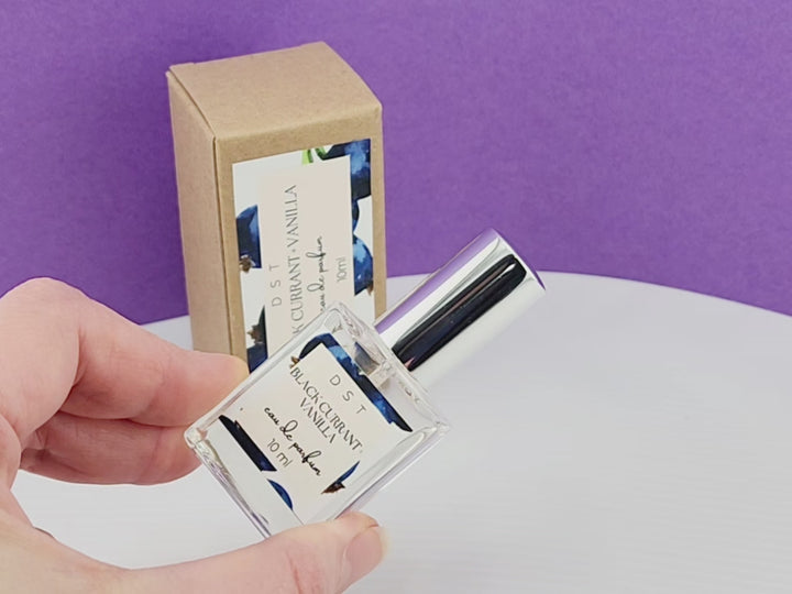 10ml bottle of Black Currant and Vanilla parfum and the box it is packaged in sitting on a white countertop with purple background. 