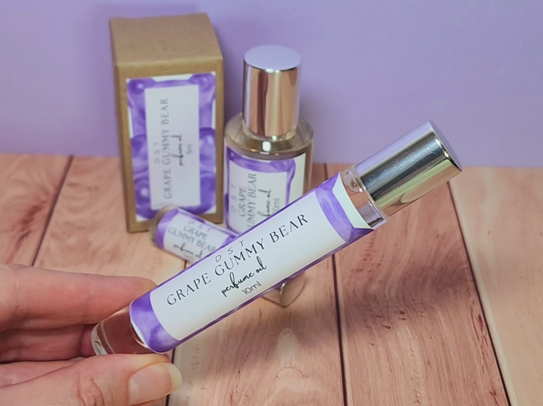 5, 10 and 30ml bottles of Grape Gummy Bear perfume oil and the box it is packaged in sitting on wood countertop with pastel purple background.